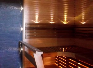 Tunnelmallinen sauna. Kiukaan takana kiviseinä. Selkänojassa ja kiukaan sivuilla ledi valot. Rakennusliike Harju-Heikkilä Oy.